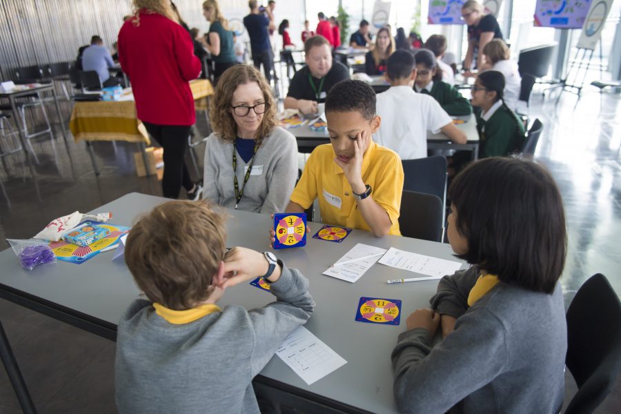 A group of children sit around a table playing the 24 Game, with their teacher watching.