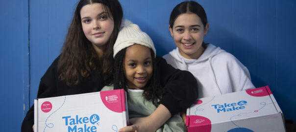 Three young Londoners smile as they hold up two Take and Make boxes.